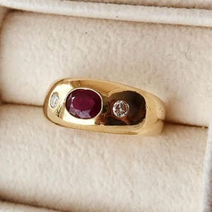 May include: A gold ring with an oval ruby and two small diamonds. The ring's band is smooth and polished. The design is classic. The ring is suitable for a special occasion.