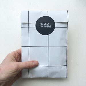 May include: White mailing envelope with a black grid pattern and a black circle sticker with the text "HELLO, I'M HERE" in white.