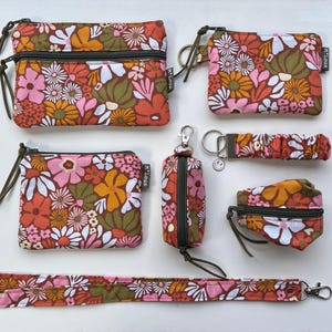 Op de afbeelding: Een verzameling accessoires met bloemenprint, waaronder etuis met ritssluiting, sleutelhangers en een lanyard. De stof heeft een retro bloemenpatroon in oranje, roze, wit en groen. Elk item heeft een donkergroene rits en trekker.