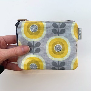 Può includere: Un piccolo portamonete con cerniera, con sfondo grigio e un motivo ripetuto di disegni floreali gialli e grigi. La borsa ha una piccola etichetta grigia con la scritta "M * Lous". La cerniera è color bronzo.