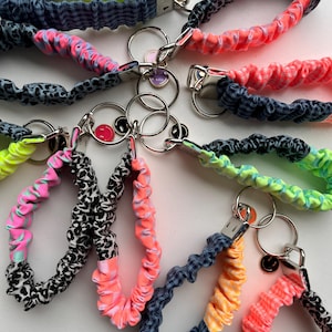 Puede incluir: Una colección de llaveros de pulsera de tela con bandas elásticas fruncidas en varios patrones y colores, incluyendo amarillo neón, rosa brillante, naranja, estampado de leopardo en blanco y negro y cuadros vichy azules, con anilla y dije.
