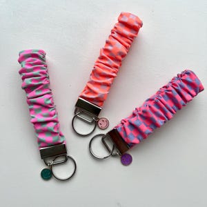 Könnte beinhalten: Drei Schlüsselanhänger mit Scrunchies in Rosa, Orange und Rosa mit Schachbrettmustern. Jeder Schlüsselanhänger hat einen silbernen Metallverschluss und einen Smiley-Anhänger. Die Scrunchies sind aus einem weichen, strukturierten Stoff.