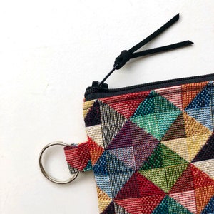 May include: A small, colorful zippered pouch with a geometric pattern. The pouch has a black zipper and a black leather strap with a loop. The pouch also has a silver ring attached to it.