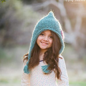 May include: A young girl wearing a teal knitted hat with a pink flower detail, a white long-sleeved shirt with colourful polka dots, and a pink tulle skirt. She is smiling and looking at the camera.