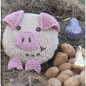 May include: A knitted pig toy with pink ears, snout, and feet. The pig is sitting on a bed of hay with a purple pear and potatoes nearby. The text "@patternervy" is visible in the upper right corner of the image.