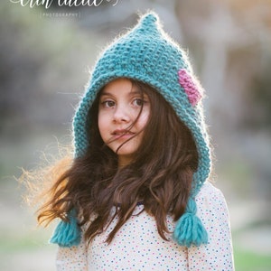 May include: A young girl wearing a teal knitted hat with a pink heart detail and a white shirt with colourful polka dots.