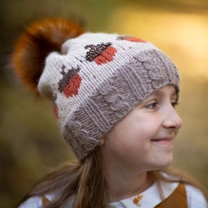 Può includere: Un bambino che indossa un cappello di lana con un pon pon marrone e un motivo a ghiande. Il cappello è bianco e marrone con accenti rossi. Il bambino sorride e guarda a destra.