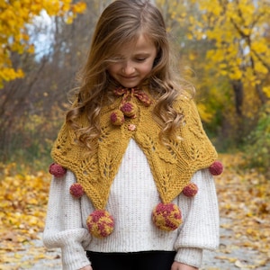 Puede incluir: Una niña pequeña lleva una chal de punto amarillo y burdeos con pompones. El chal tiene un patrón de encaje y se lleva sobre un suéter blanco. La niña está de pie en un bosque con follaje otoñal.