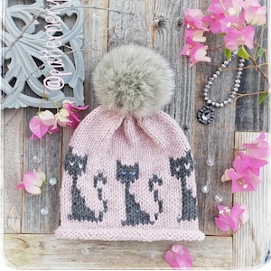 May include: A pink knitted hat with a grey pom pom and a black cat design. The hat is on a wooden background with pink flowers.