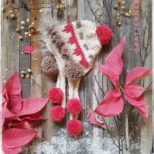 May include: A white and brown knitted hat with a pink pom-pom on top and a faux fur trim. The hat has a long scarf with pink pom-poms attached.