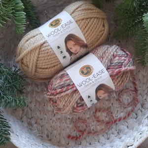 May include: Two skeins of Lion Brand Wool-Ease yarn. One skein is a beige color and the other is a multi-colored skein with red, white, and brown yarn. The yarn is in a white wicker basket.