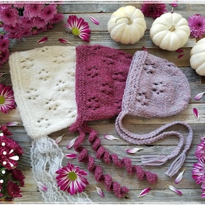 May include: Two knitted baby bonnets with lace trim, one white with a delicate pattern and one pink with a more intricate design. The bonnets are displayed on a wooden surface with white pumpkins and pink flowers.
