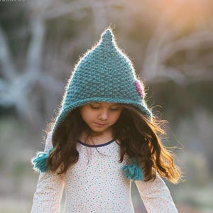 Op de afbeelding: Een jong meisje draagt ​​een turquoise gebreide muts met een roze bloem, een wit lang mouw shirt met een bloemenpatroon, een roze tule rok en roze laarzen. Ze staat in een grasveld met een wazige achtergrond.