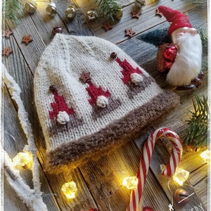 May include: A white knitted hat with a brown brim and a pattern of red and white Christmas trees. The hat is decorated with wooden buttons and stars. The hat is surrounded by Christmas decorations, including a candy cane, bells, and lights.