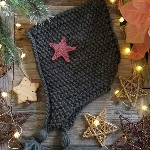 May include: A dark grey knitted hat with a pink star and two tassels. The hat is on a wooden surface with fairy lights and decorative stars.