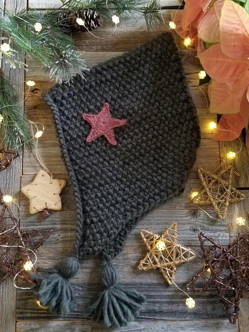 May include: A dark grey knitted hat with a pink star and two tassels. The hat is on a wooden surface with fairy lights and decorative stars.