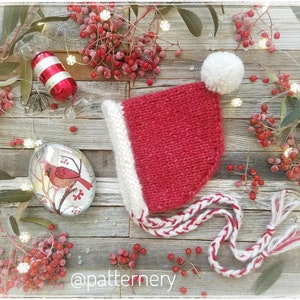 Könnte beinhalten: Eine rote gestrickte Weihnachtsmütze mit einem weißen Bommel und weißem Besatz. Die Mütze hat eine lange weiße und rote geflochtene Schnur mit Quasten. Die Mütze liegt auf einer Holzoberfläche mit roten Beeren und einem Glasornament mit einem Kardinal.