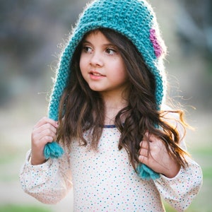 May include: A young girl wearing a teal knitted hat with a pink flower accent. The hat has a long pointed tip and a pom-pom on the end. She is wearing a white dress with colourful polka dots and a pink skirt.