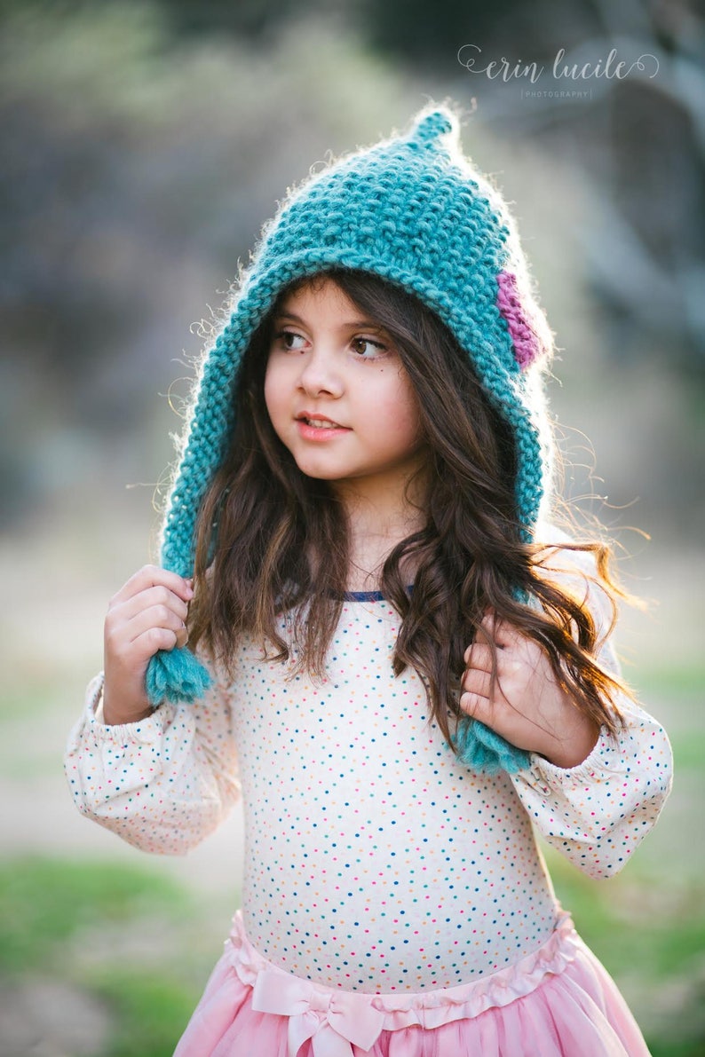 May include: A young girl wearing a teal knitted hat with a pink flower accent. The hat has a long pointed tip and a pom-pom on the end. She is wearing a white dress with colourful polka dots and a pink skirt.