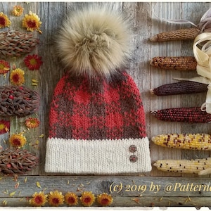 May include: A red and brown plaid knitted hat with a white brim and a fluffy brown pom pom. The hat is on a wooden background with dried corn and flowers.