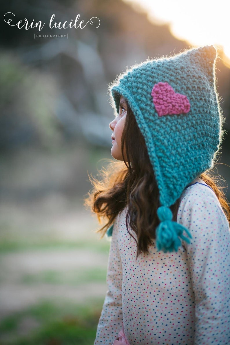 May include: A young girl wearing a teal blue knitted hat with a pink heart on the front. The hat has a long pointed hood and a fluffy pom-pom on the end. The girl is wearing a white shirt with colourful polka dots and is looking up at the sky.