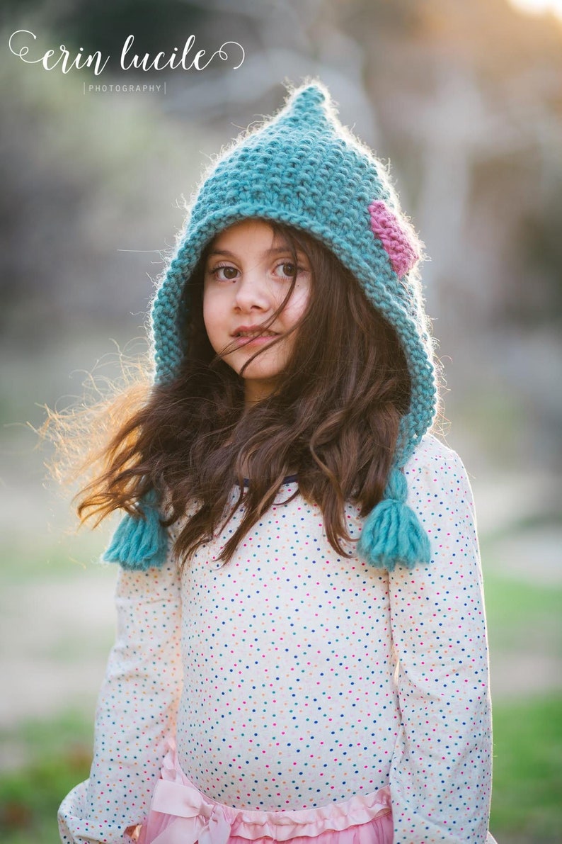 May include: A young girl wearing a teal knitted hat with a pink heart detail and a white shirt with colourful polka dots.