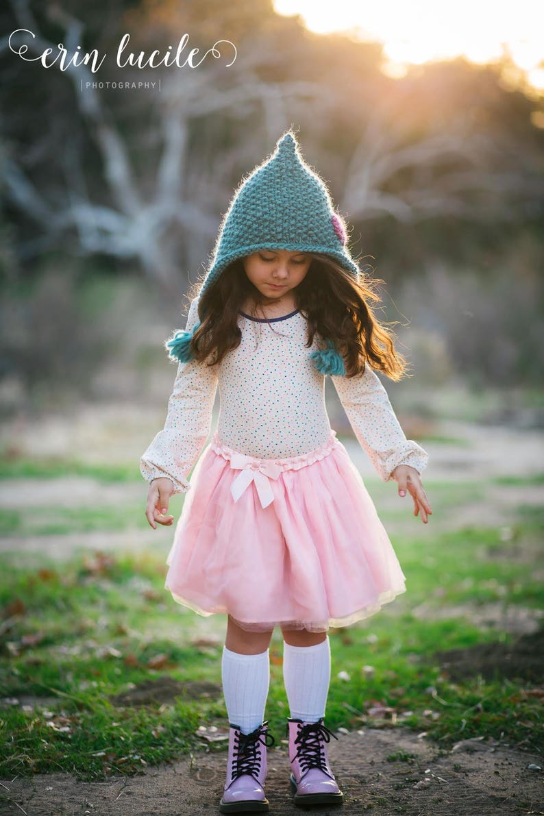 May include: A young girl wearing a teal knitted hat with a pink flower, a white long-sleeved shirt with a floral pattern, a pink tulle skirt, and pink boots. She is standing in a grassy field with a blurred background.
