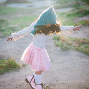 May include: A young girl wearing a teal knitted hooded hat, a pink tulle skirt, and pink boots with black laces. She is standing on a dirt path with green grass on either side.