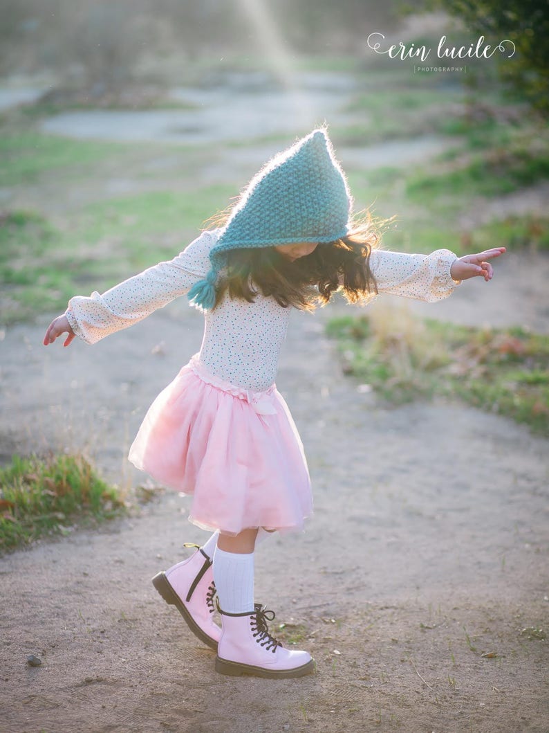 May include: A young girl wearing a teal knitted hooded hat, a pink tulle skirt, and pink boots with black laces. She is standing on a dirt path with green grass on either side.