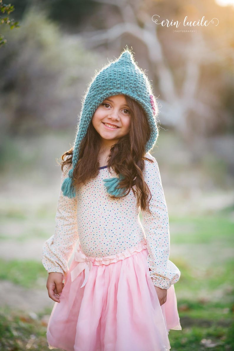 May include: A young girl wearing a teal knitted hat with a pink flower detail, a white long-sleeved shirt with colourful polka dots, and a pink tulle skirt. She is smiling and looking at the camera.
