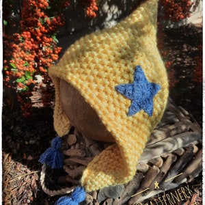 May include: A yellow knitted baby hat with a blue star and blue tassels. The hat is sitting on a piece of driftwood with the text "@patternervy" on it.