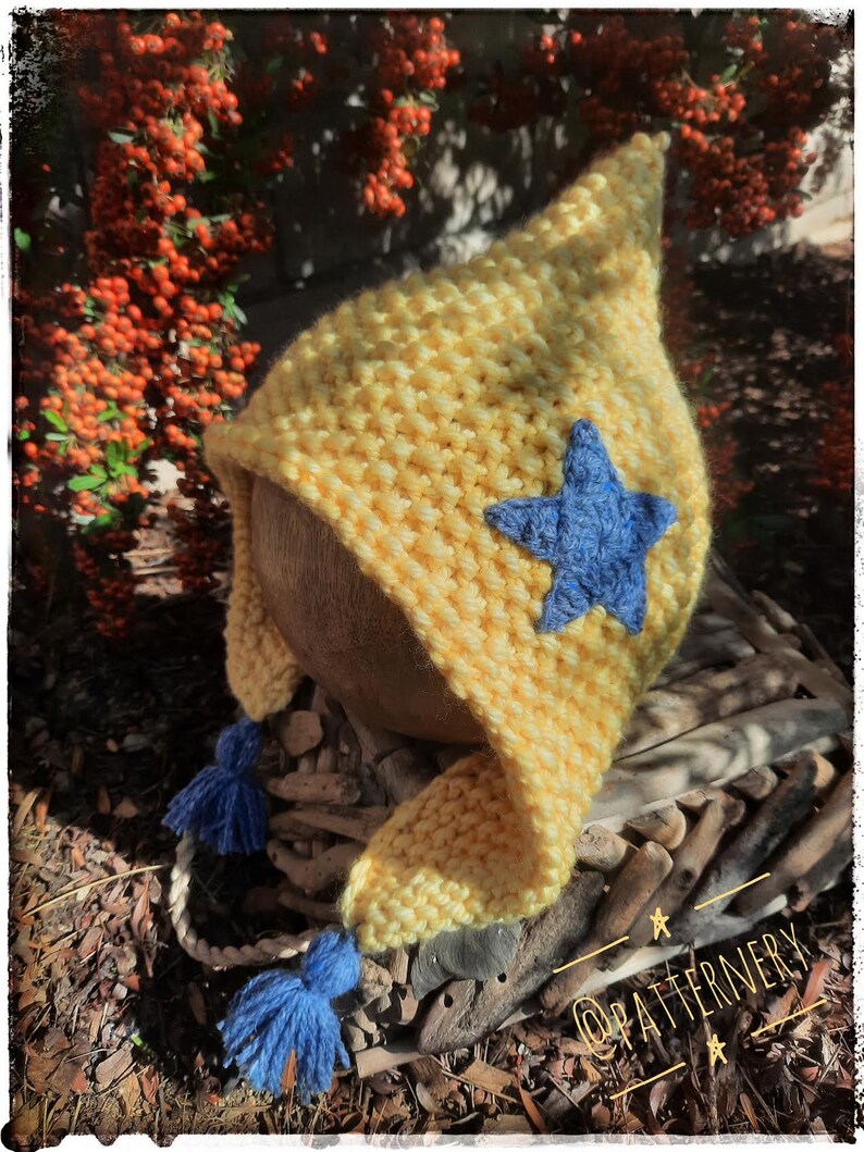 May include: A yellow knitted baby hat with a blue star and blue tassels. The hat is sitting on a piece of driftwood with the text "@patternervy" on it.