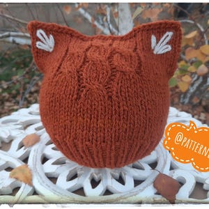May include: A rust-colored knitted hat shaped like a fox's head, featuring pointed ears with white embroidered details. The hat has a textured knit pattern and is displayed on a white, ornate surface. The image includes the text "@PATTERNRY".