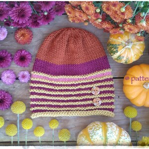 May include: A knitted hat with a striped pattern in orange, pink, and yellow with two buttons. The hat is surrounded by autumn-coloured flowers and pumpkins.