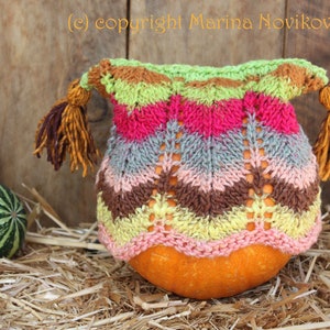 May include: A colorful knitted pumpkin cozy with a chevron pattern in shades of pink, green, brown, and yellow. The cozy has two tassels and is placed over an orange pumpkin. The background includes hay and gourds.