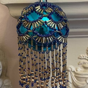 May include: A teal Christmas bauble with a beaded design. The bauble is covered in a pattern of navy blue and gold-coloured beads. Long strands of gold and navy blue beads hang from the bottom of the bauble.