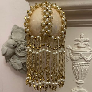 May include: Cream-coloured Christmas bauble with gold and pearl accents. The spherical ornament is decorated with gold beads and faux pearls. Long strands of pearls and gold beads hang from the bottom, creating a decorative fringe.
