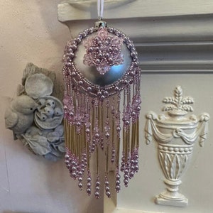 May include: A silver Christmas bauble with a pink beaded design. The bauble has a beaded fringe and a pink flower design on the front.