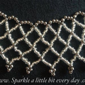 Könnte beinhalten: Eine silberne Perlenkette mit geometrischem Muster. Die Kette besteht aus kleinen, runden Perlen, die in einem Diamantenmuster zusammengefügt sind. Die Perlen sind silberfarben und haben eine glänzende Oberfläche.