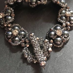 Può includere: Un braccialetto di perline grigie con un design a fiore e accenti di cristallo.