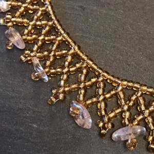 May include: Close-up of a gold beaded necklace with a geometric design. The necklace features small, round gold beads and clear, teardrop-shaped accents. The intricate pattern and delicate details create a sophisticated look.