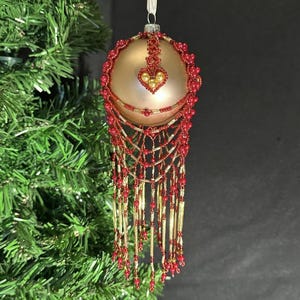 May include: A gold Christmas ornament with red beaded accents and a heart-shaped design. The ornament has a beaded fringe and is hanging from a white ribbon. The ornament is hanging in front of a green Christmas tree.