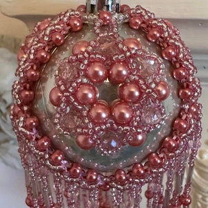 May include: A decorative Christmas ornament with a pearlescent finish, adorned with pink beads and faux pearls. The ornament features a beaded floral design and dangling beaded fringe, creating a festive and elegant appearance.