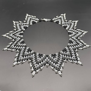 May include: A striking beaded necklace in black, white, and silver. The necklace features a geometric design with a sunburst or star-like shape. The intricate beadwork creates a bold, eye-catching accessory, perfect for adding a touch of elegance to any outfit.