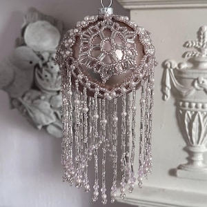 Peut inclure: Une décoration de Noël en forme de boule, avec une base sphérique rose pâle. L'ornement est orné de perles argentées et d'une frange de perles. Le dessus présente un motif géométrique détaillé. Une boucle en métal argenté est en haut pour suspendre.