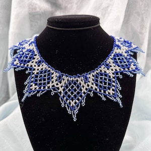 May include: A beaded necklace in shades of blue and white, displayed on a black mannequin. The necklace features a geometric design with a scalloped edge. The beads are intricately woven, creating a delicate and detailed piece of jewelry.