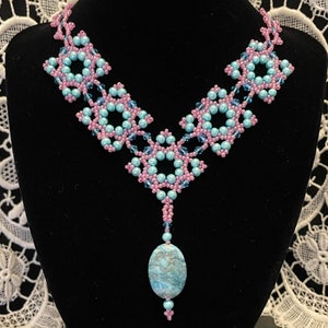 Può includere: Una collana di perline turchesi e rosa con un ciondolo in pietra turchese. La collana è realizzata con perline piccole e ha un design delicato e intricato.