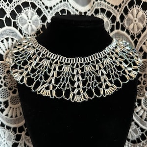 May include: A silver beaded choker necklace with a scalloped design. The necklace is displayed on a black velvet stand.