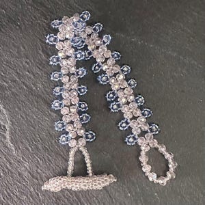Pode incluir: Uma pulseira de contas com um design delicado. A pulseira apresenta contas transparentes e azuis dispostas em um padrão floral. A pulseira é exibida em uma superfície cinza escuro.