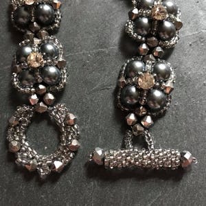 May include: A handmade bracelet with a toggle clasp, featuring dark grey pearl beads, silver seed beads, and faceted silver beads. Each of the three floral-shaped sections has a clear, faceted crystal at its centre. The bracelet is shown close-up.